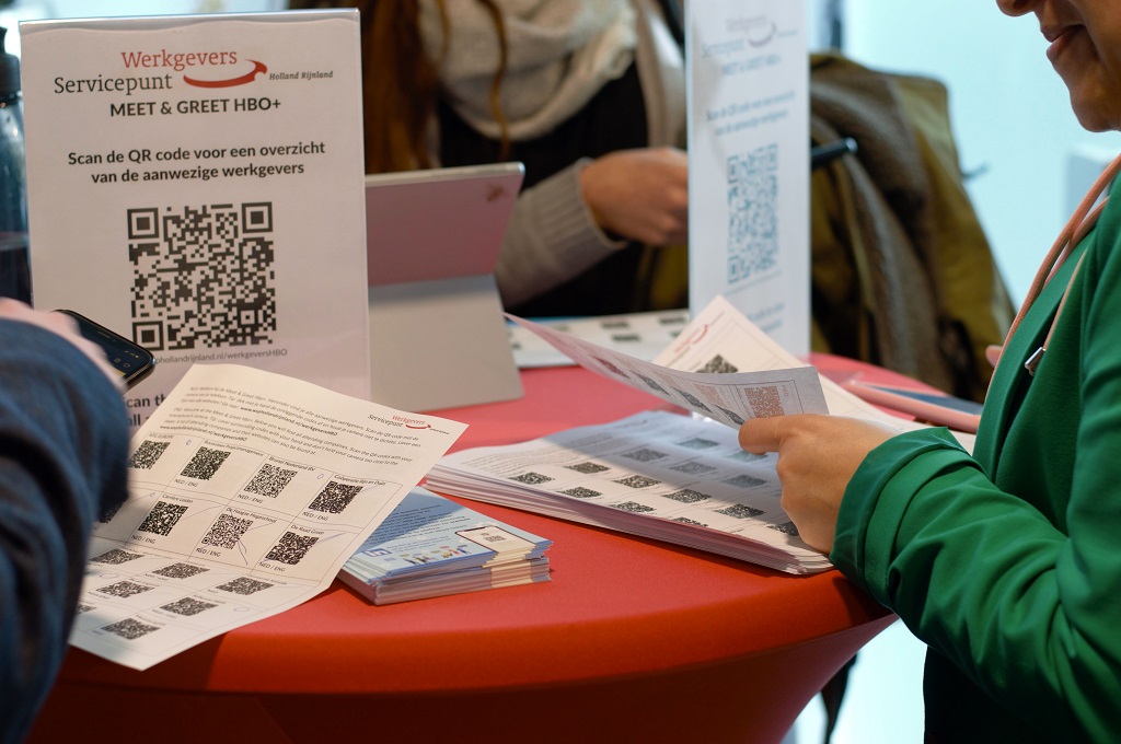 Een close-up van een sta-tafel. Je ziet mensen een lijst met QR codes bekijken en met hun telefoon de codes scannen. Op de papieren die op tafel liggen staan tips over netwerken via LinkedIn.