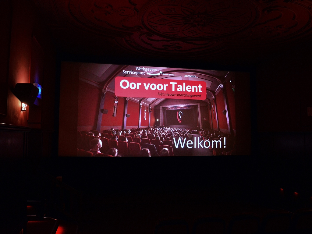 Je ziet een donkere bioscoopzaal. Op het scherm staat een foto van een klassiek uitziende bioscoopzaal, met de tekst "Welkom" en "Oor voor Talent".