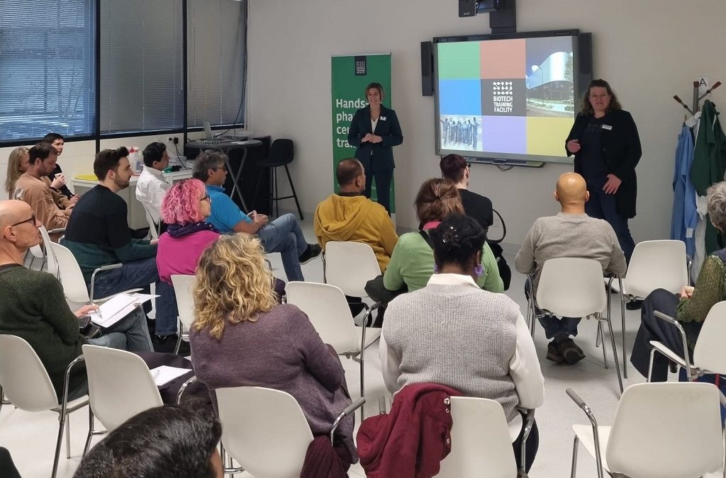 Een groep mensen zit met hun gezicht van de camera af. Ze kijken naar de presentatie van twee vrouwen. Op het scherm staat een kleurrijke plaat met "Biotech Training Facility" in het midden.