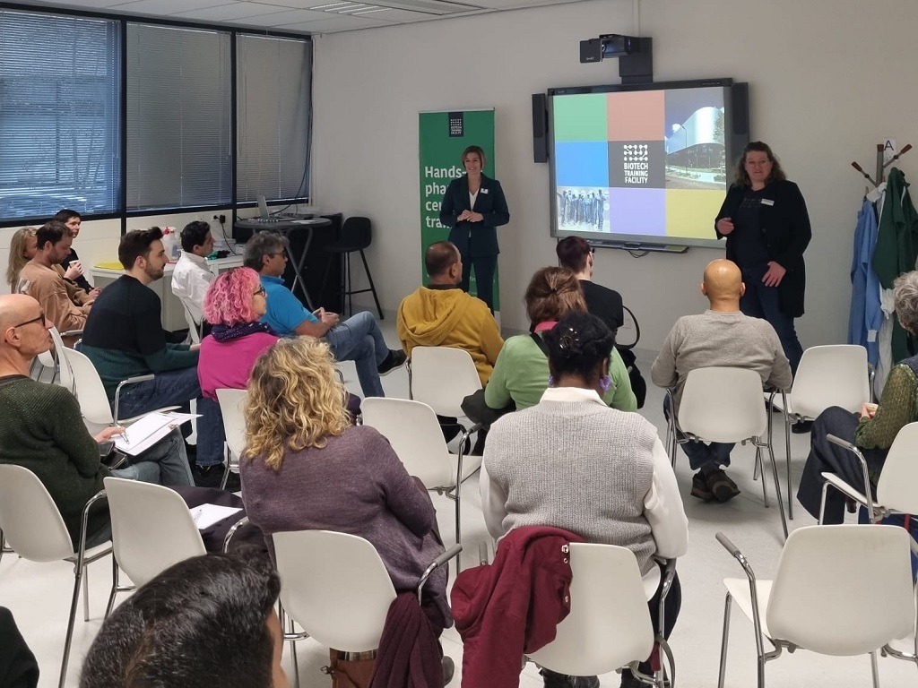 Een groep mensen zit met hun gezicht van de camera af. Ze kijken naar de presentatie van twee vrouwen. Op het scherm staat een kleurrijke plaat met "Biotech Training Facility" in het midden.